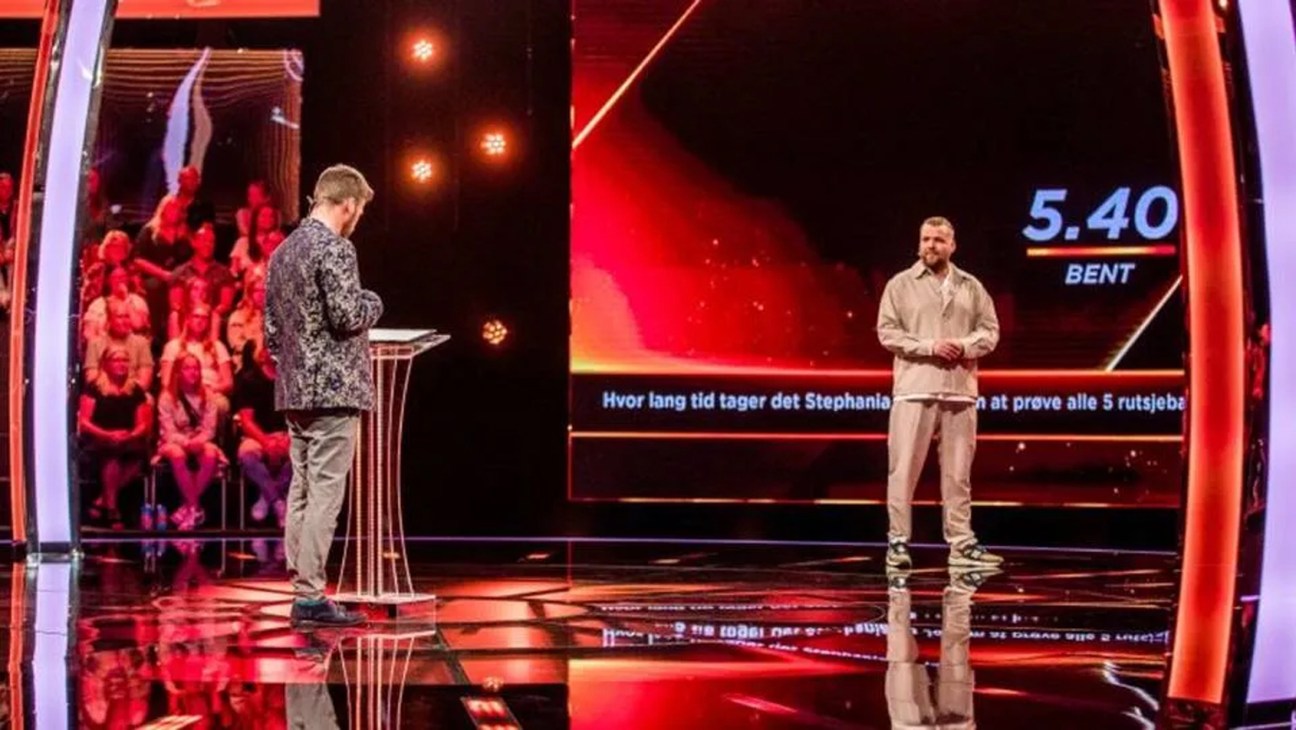 Two men on a brightly lit TV game show set with red neon lighting and an audience in the background.