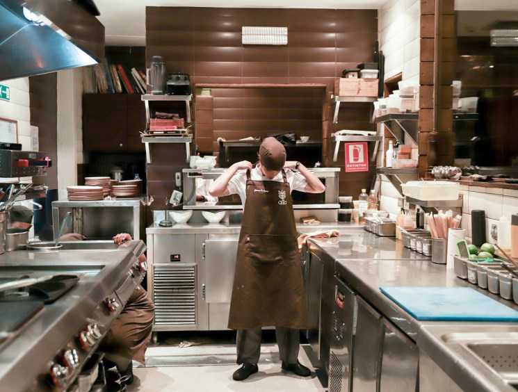 A chef in a brown apron stands in the center of a busy professional kitchen surrounded by stainless steel equipment and prep stations.