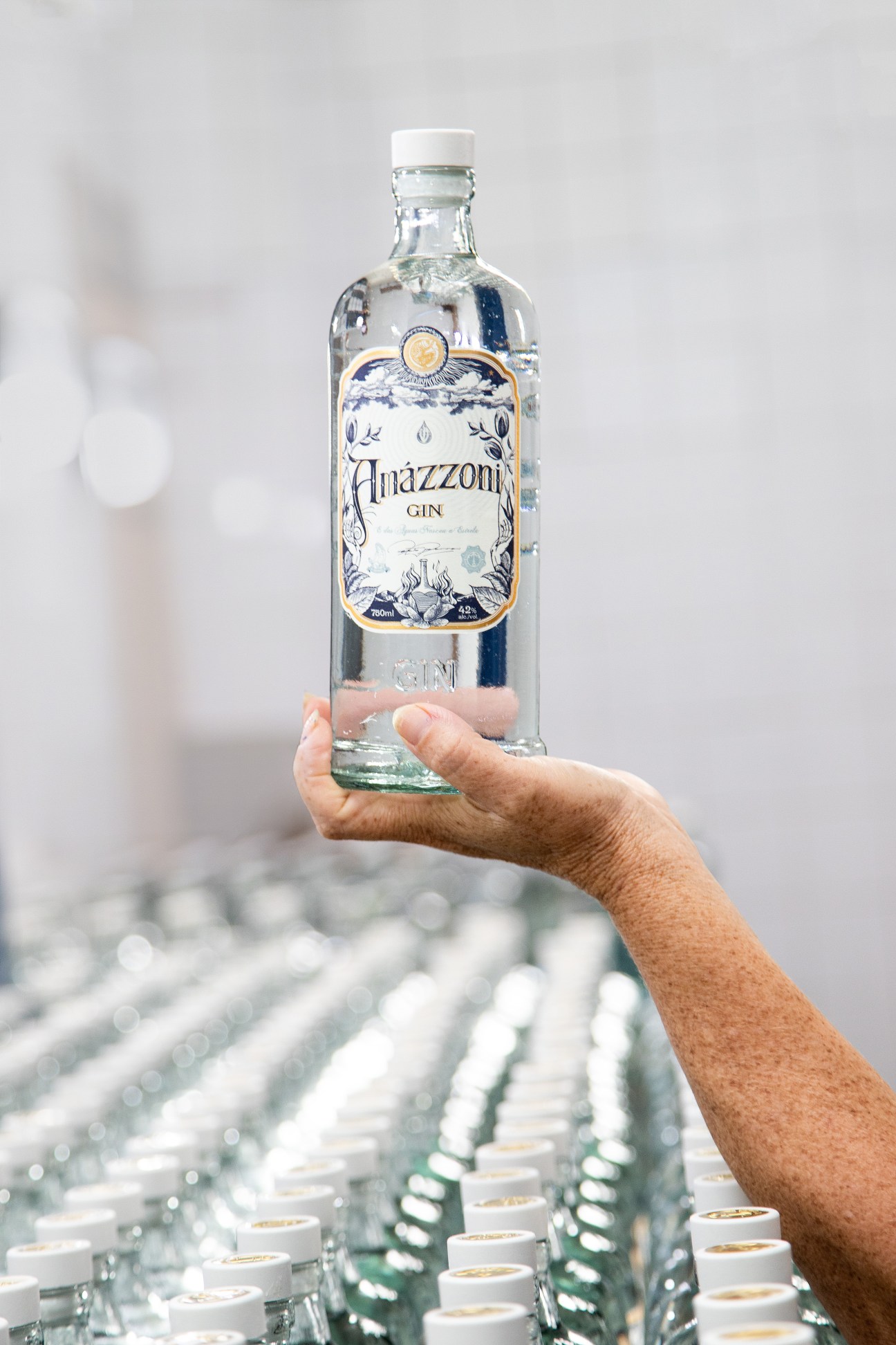 Hand holding bottle of Amázzoni gin with distillery bottles visible in blurred background