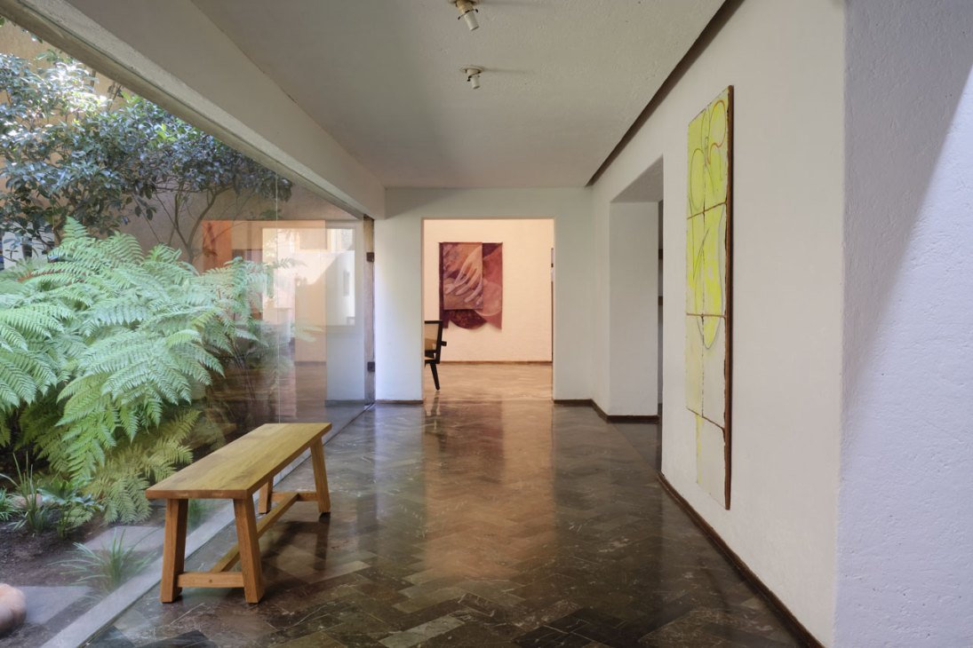 Modern gallery interior with concrete floors, white walls displaying colorful artworks, wooden bench, and garden courtyard