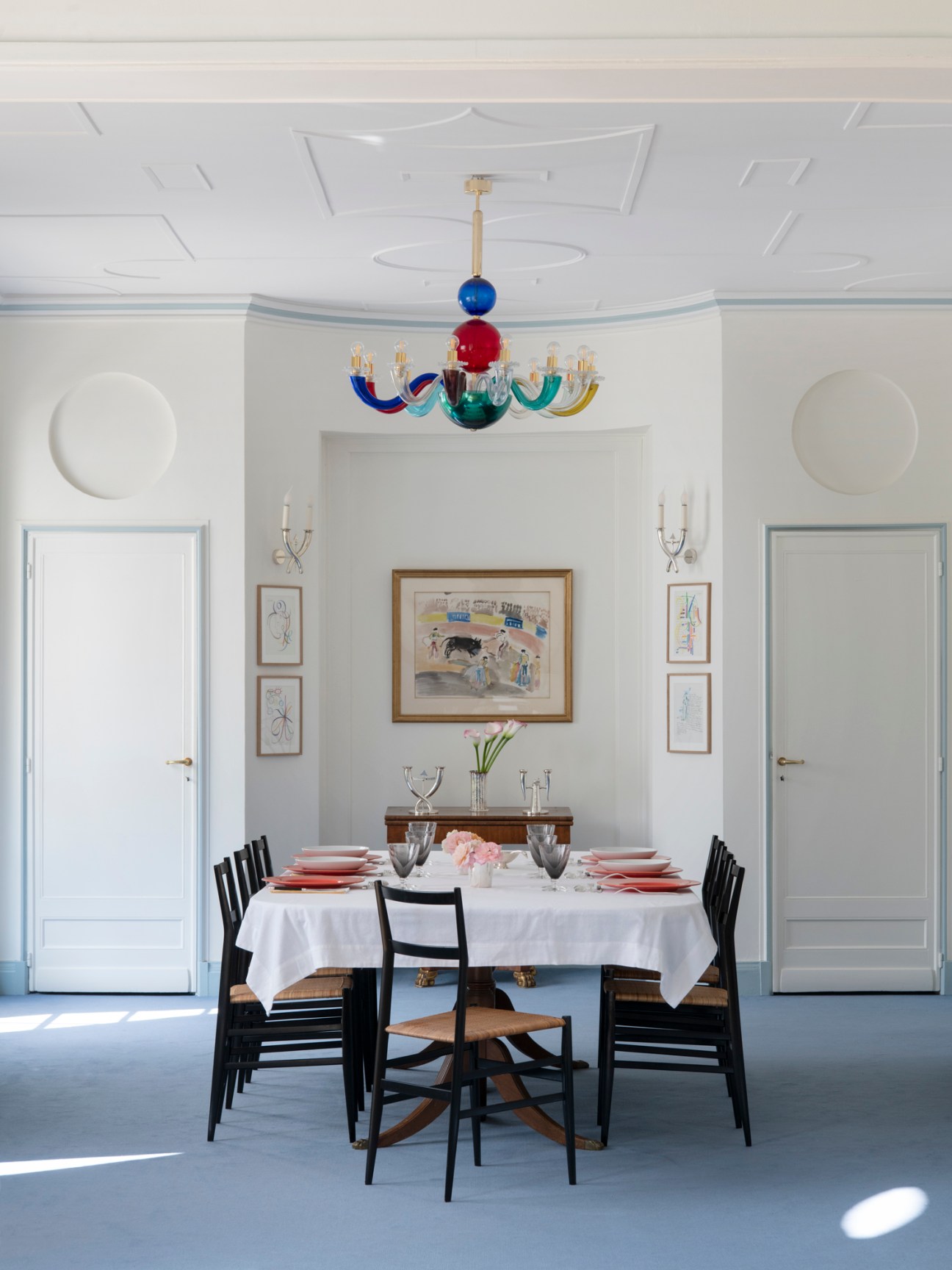 Dining room with white tablecloth, black chairs, colorful glass chandelier and framed artwork on white walls