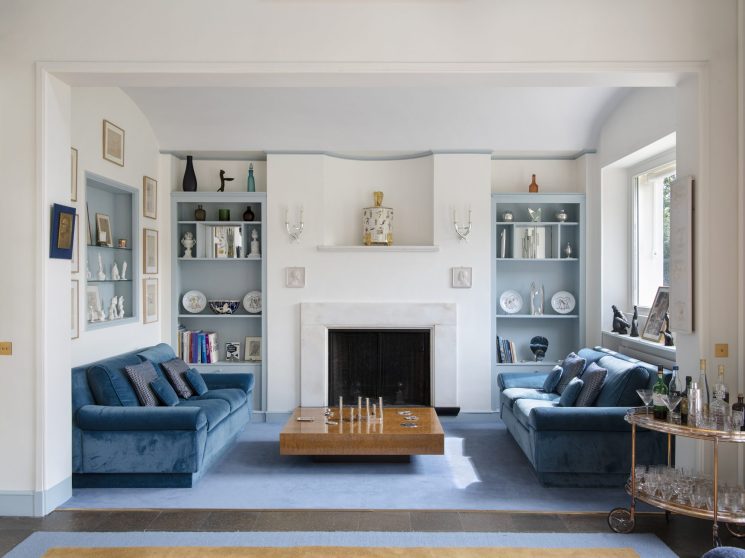 Elegant living room with blue velvet sofas, white fireplace, built-in shelving, and modern furnishings