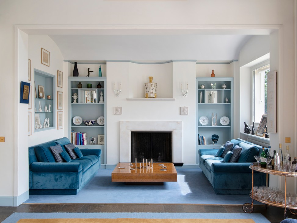 Elegant living room with blue velvet sofas, white fireplace, built-in shelving, and modern furnishings