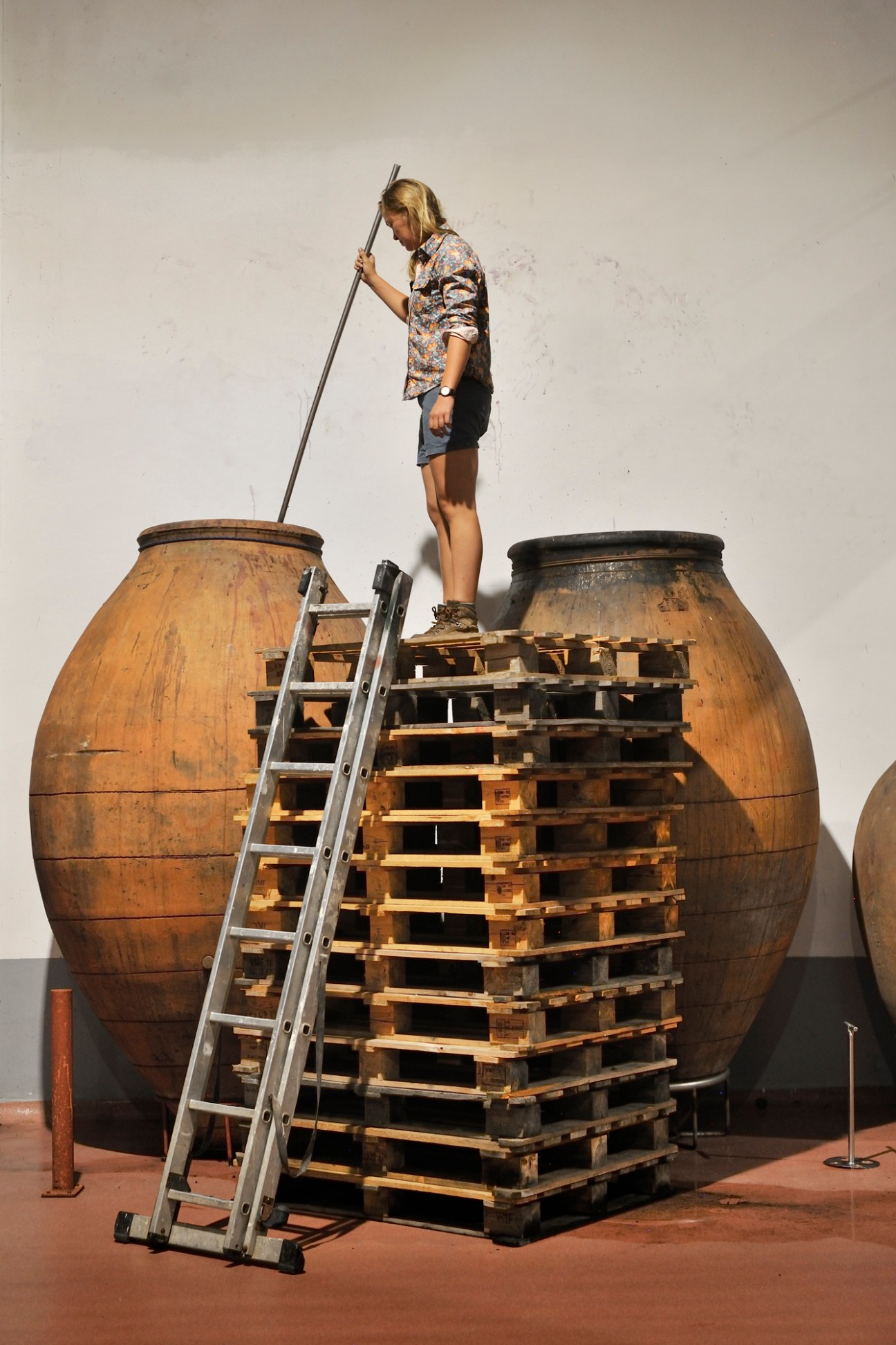 Anna Kristina Jørgensen standing on wooden pallets using a long tool to stir wine in large clay amphora vessels