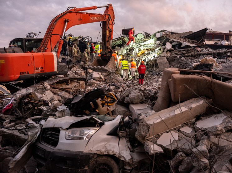 Rescue workers in high-visibility vests search through earthquake rubble with an orange excavator amid destroyed buildings