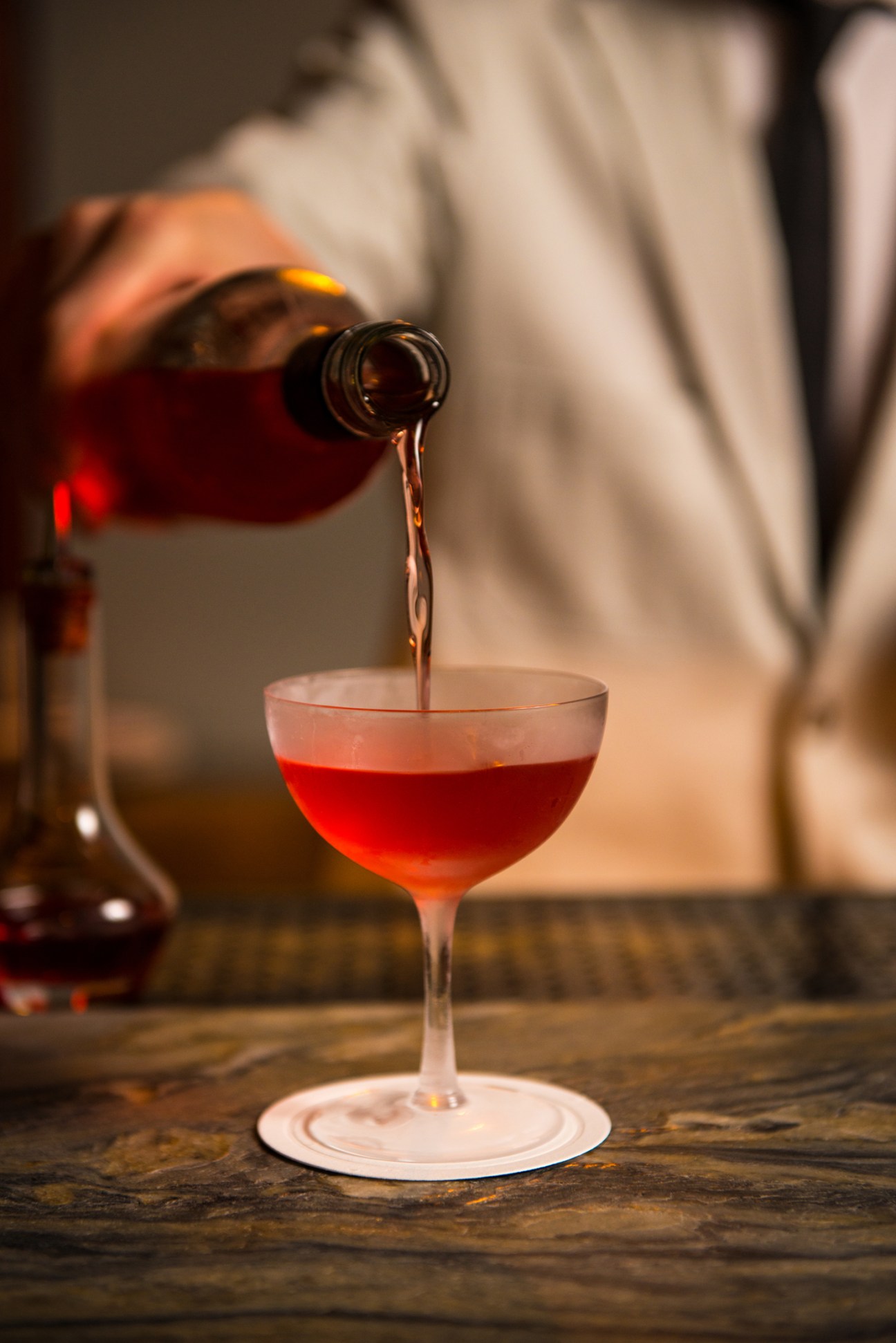 Red cocktail being poured from bottle into coupe glass on wooden bar surface with warm ambient lighting