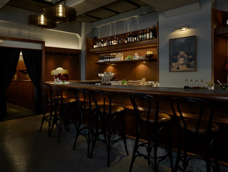 Dimly lit bar interior with dark wooden counter, black metal stools, bottles on shelves, and warm ambient lighting