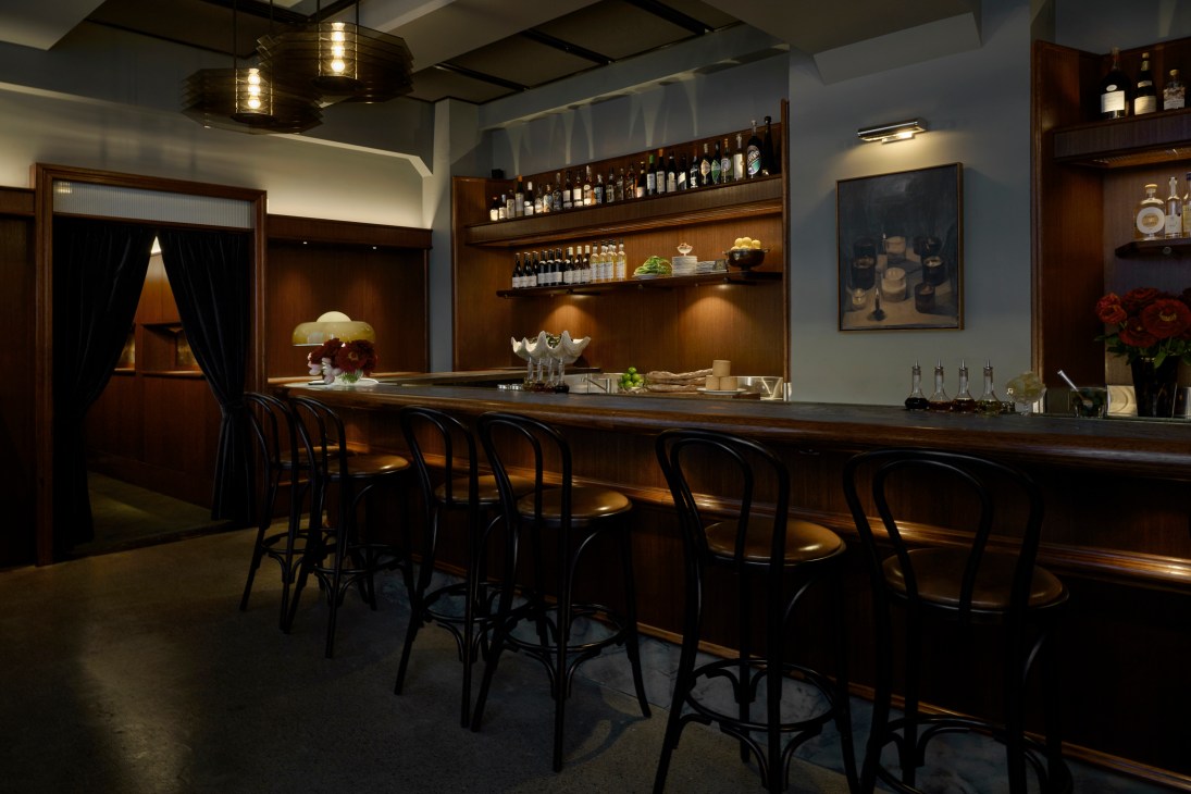Dimly lit bar interior with dark wooden counter, black metal stools, bottles on shelves, and warm ambient lighting