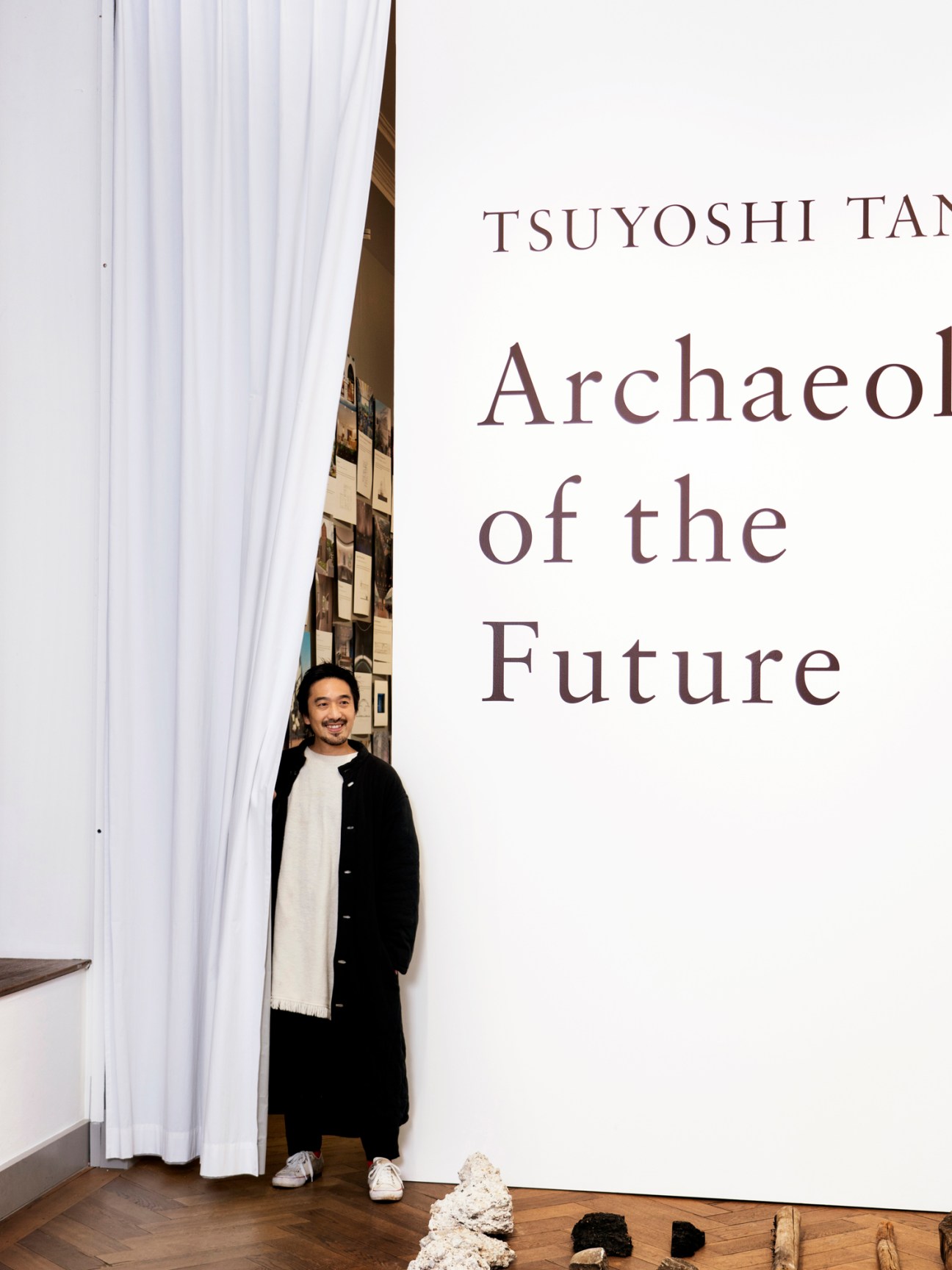Architect Tsuyoshi Tane standing beside white curtain at his exhibition featuring large text reading Archaeology of the Future