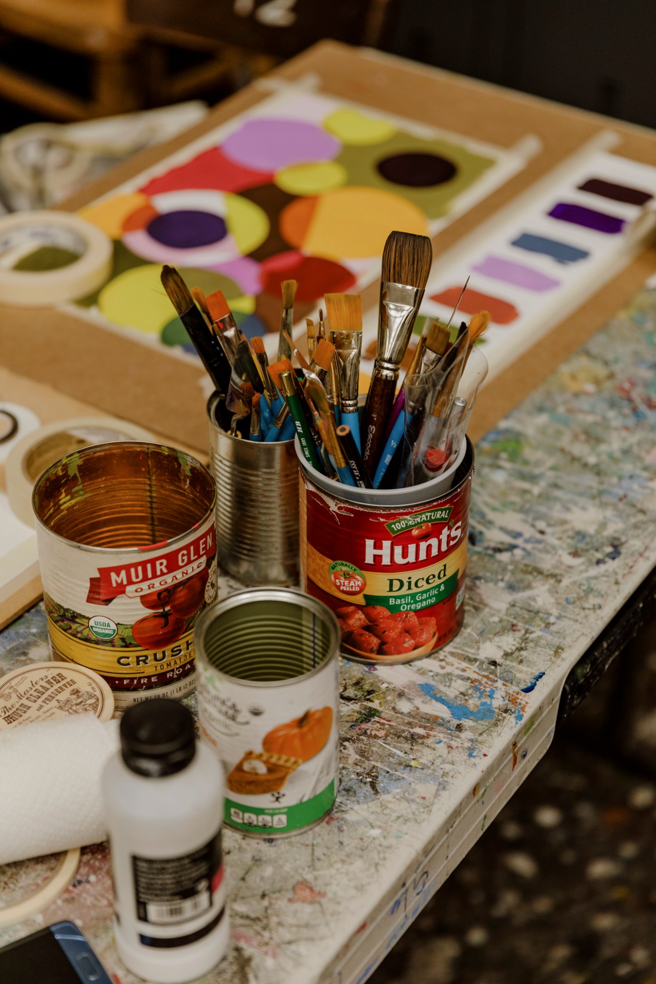Art supplies including paint brushes in tin cans, paint tubes, and food cans repurposed as brush holders on a paint-splattered work surface.