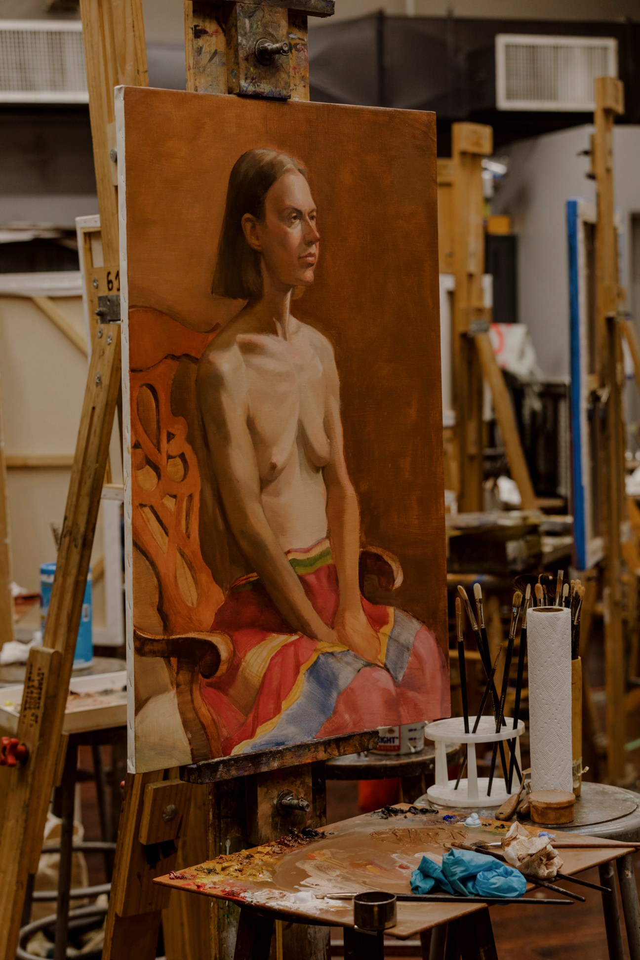 Portrait painting of a seated woman with long dark hair wearing red draped fabric against a brown background, displayed on an easel in an artist's studio.