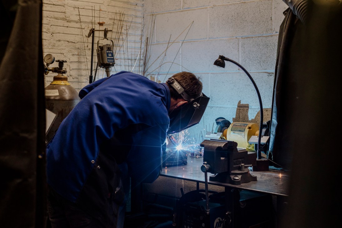 A welder in blue protective gear works at a metal sculpture studio with bright sparks flying from the welding torch.