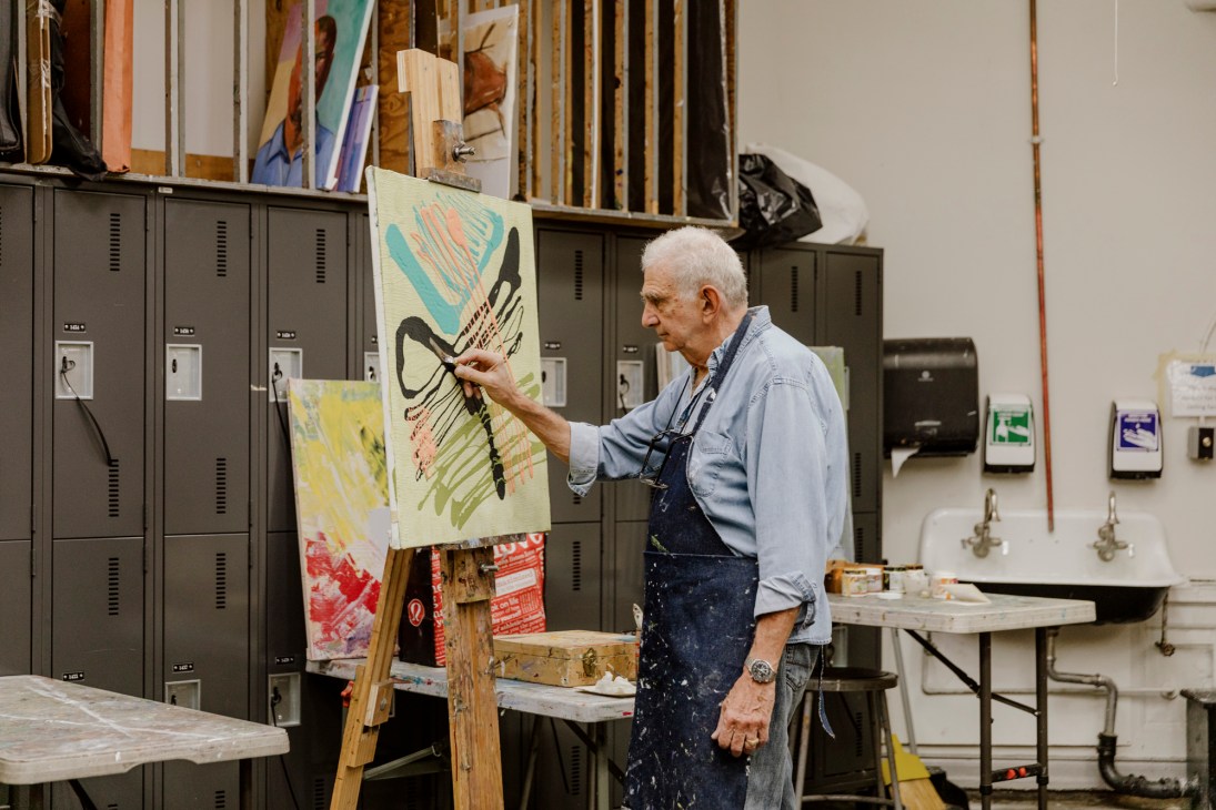 An elderly man in a light blue shirt and paint-splattered apron works on an abstract painting in an art studio.