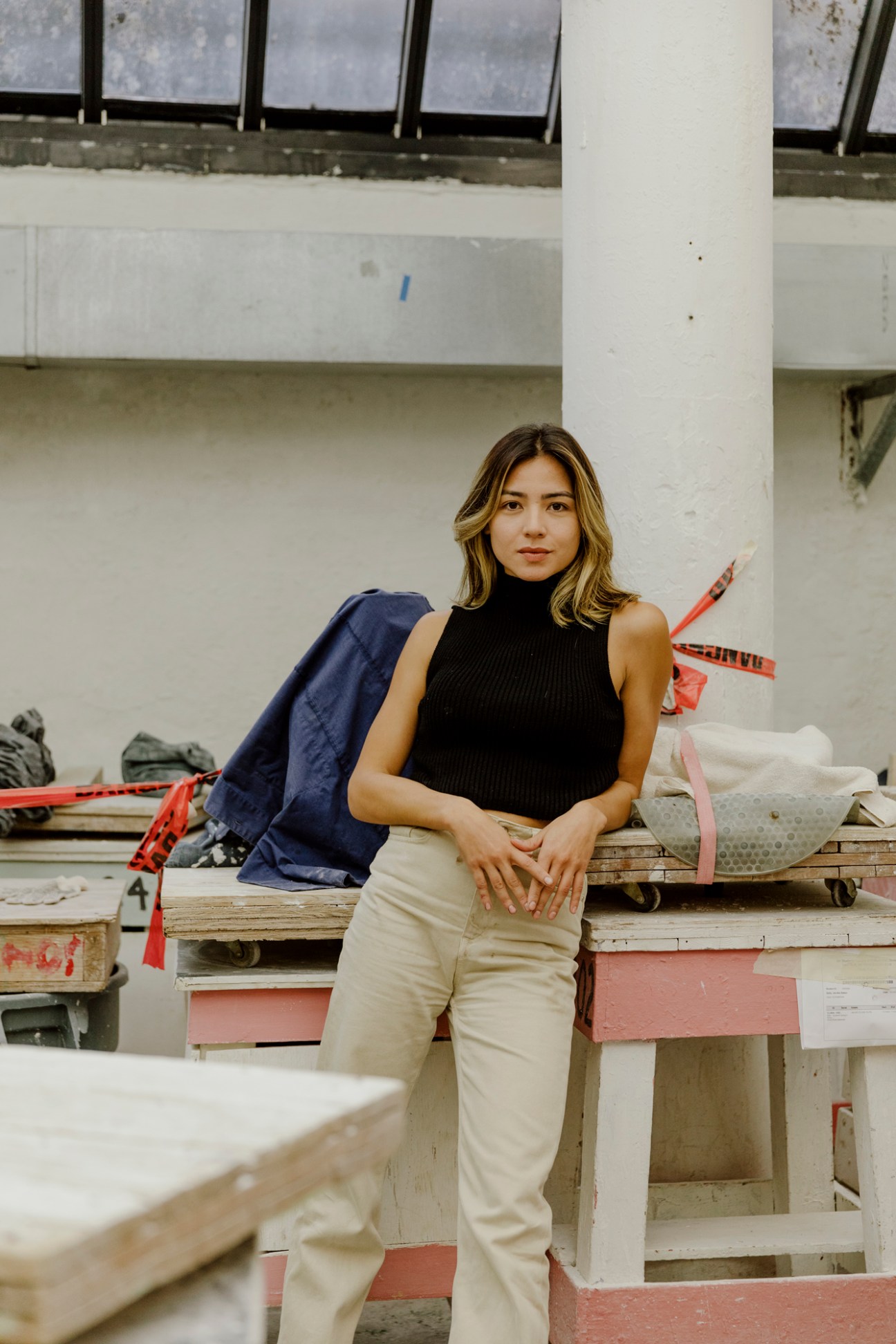 Woman in black sleeveless turtleneck and cream pants standing in art studio with fabric and materials.