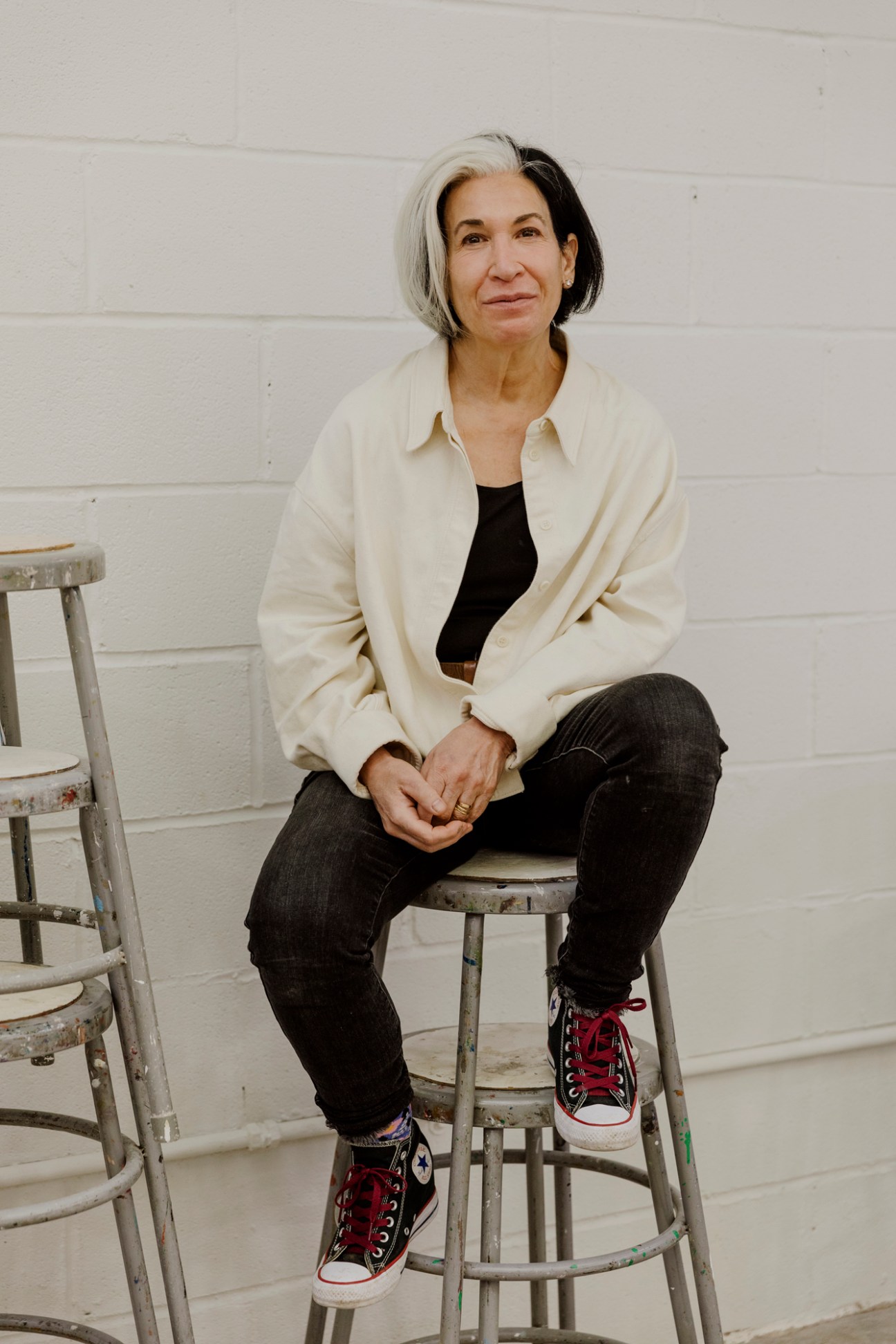 Portraitist and teacher Sharon Sprung sits on a stool against a white brick wall, wearing a cream jacket over black clothing and red sneakers.