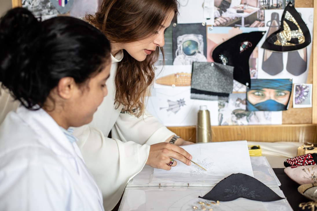 Two women examine design sketches and fabric samples at a work table in a studio.