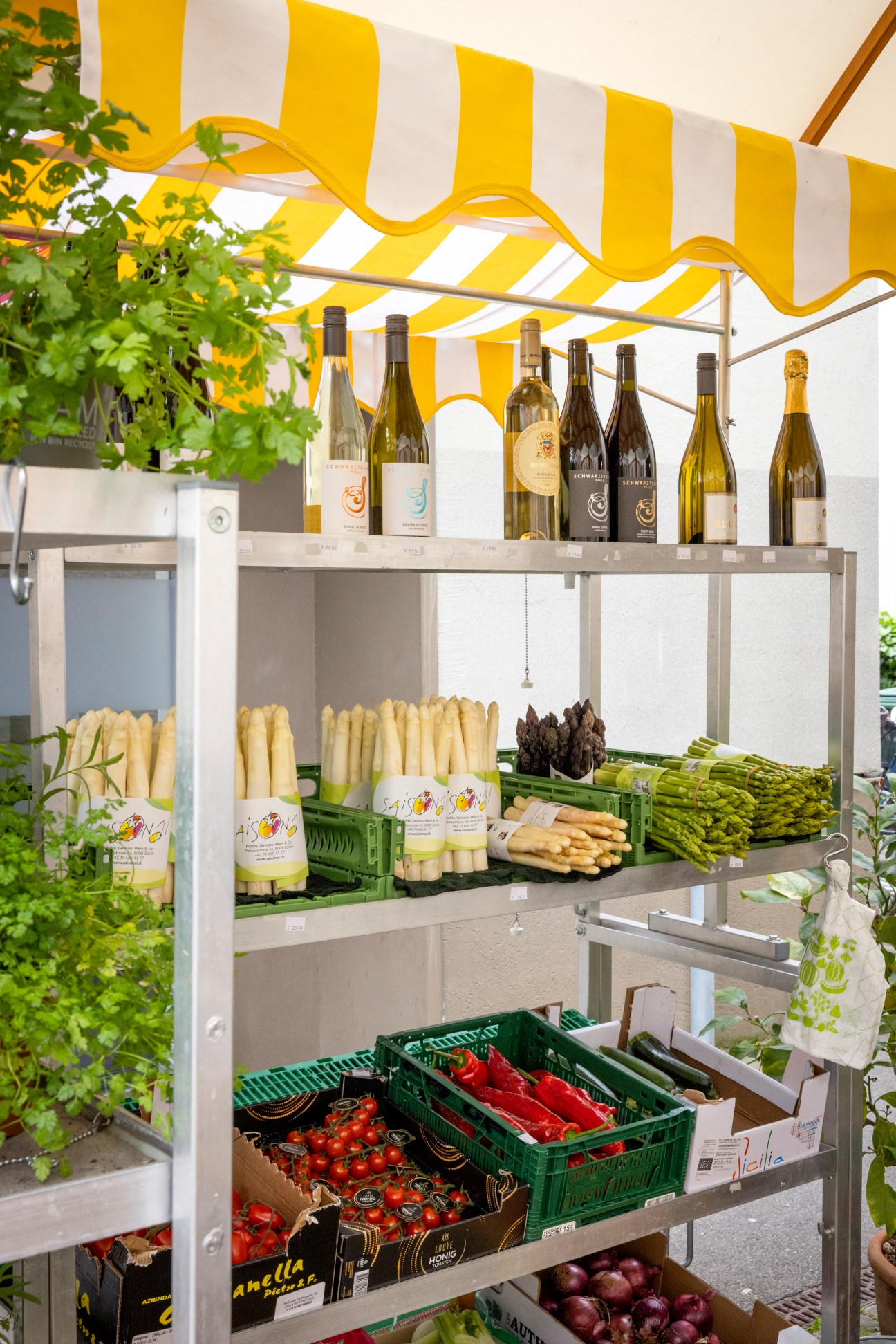 Fresh vegetables displayed on metal shelving unit under striped yellow and white awning
