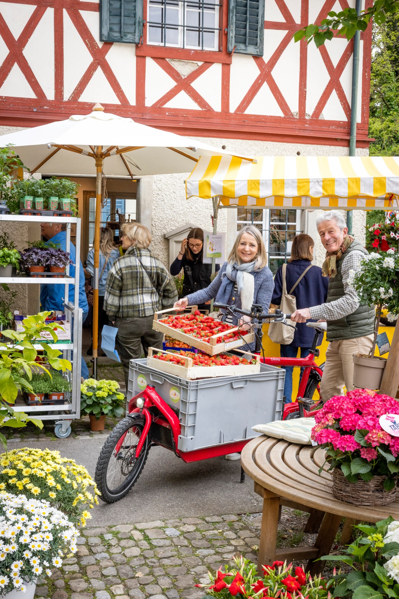 Customers shopping at an outdoor market with red bicycle cart, flowers and traditional half-timbered building