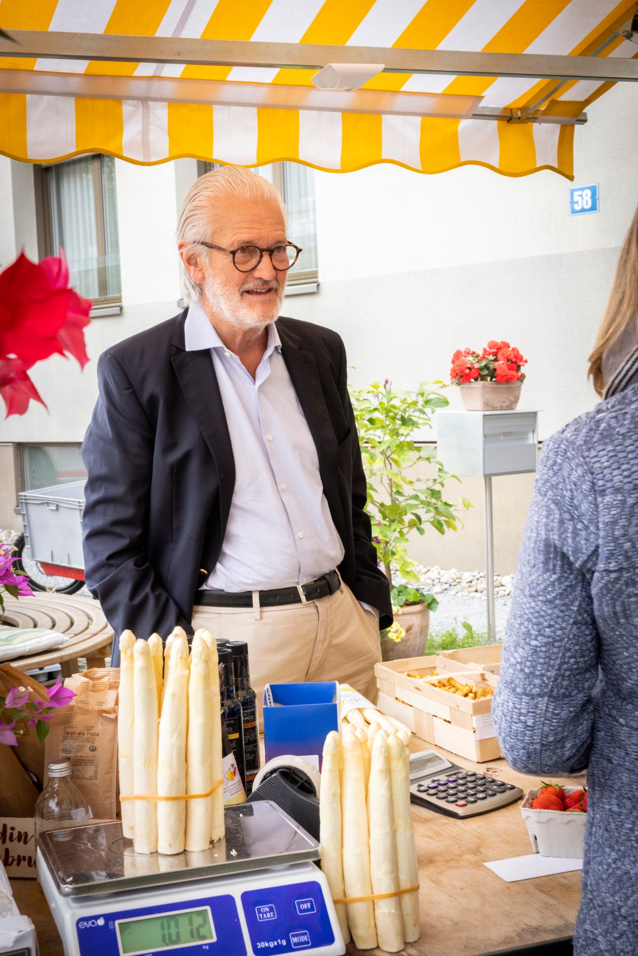 Man in glasses and dark blazer standing behind counter with white asparagus bundles and calculator at specialty food shop