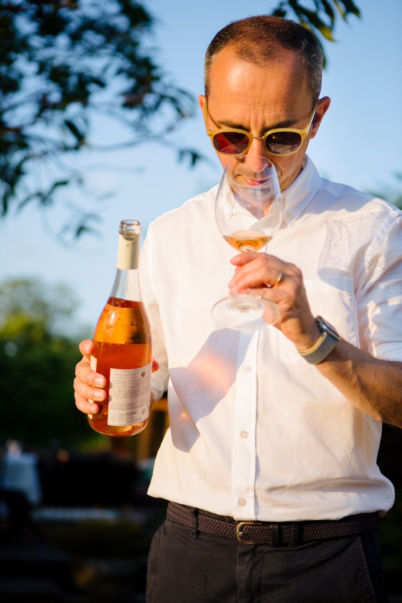 Nuno Guedes Vaz Pires holds a wine bottle and glass, examining an Azorean rosé outdoors in sunlight