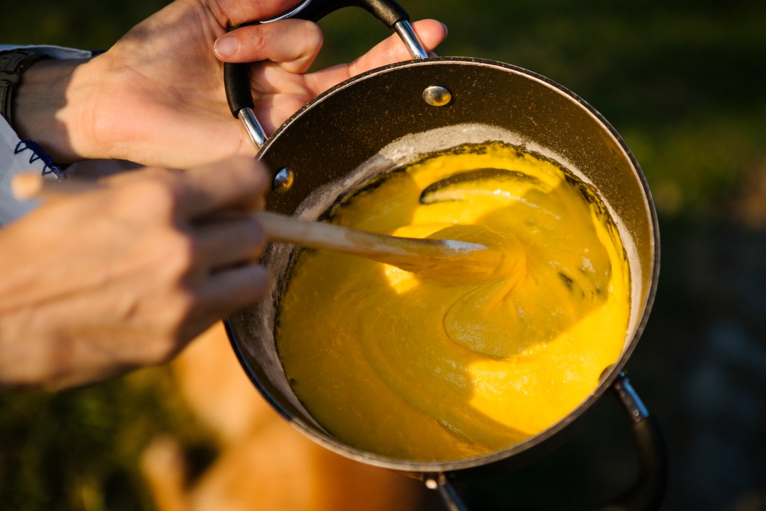 Hands stirring golden yellow custard mixture in a saucepan with a wooden spoon