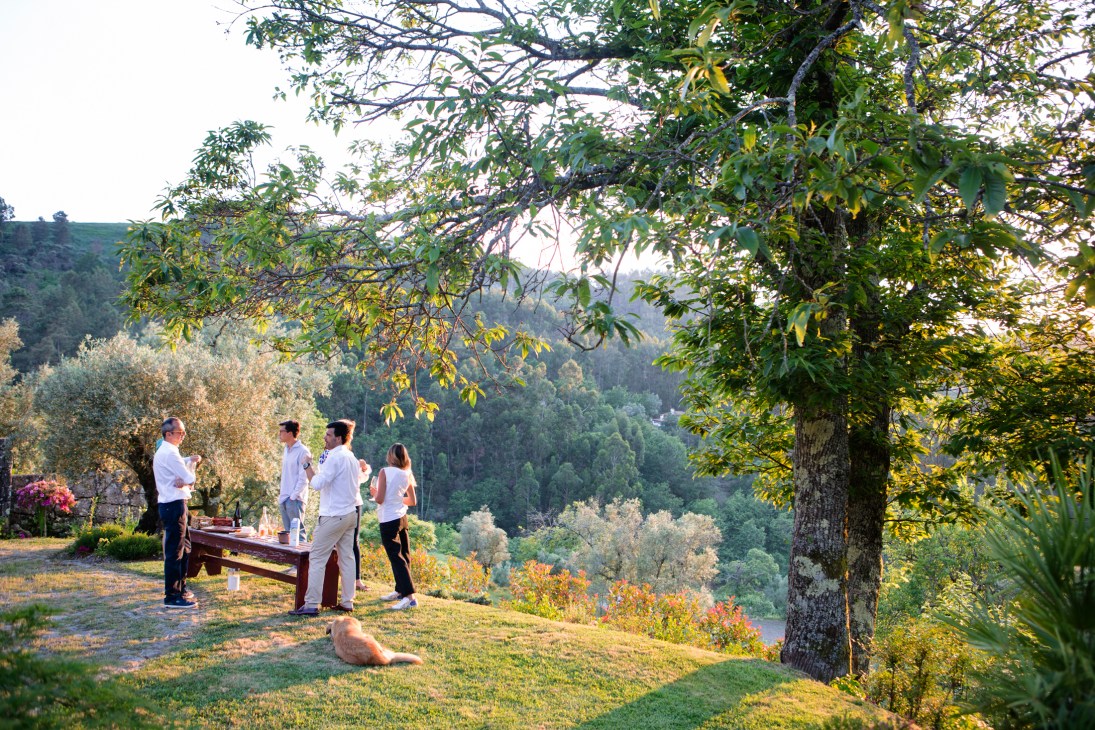Group of people gathered around outdoor table in garden setting with rolling hills in background