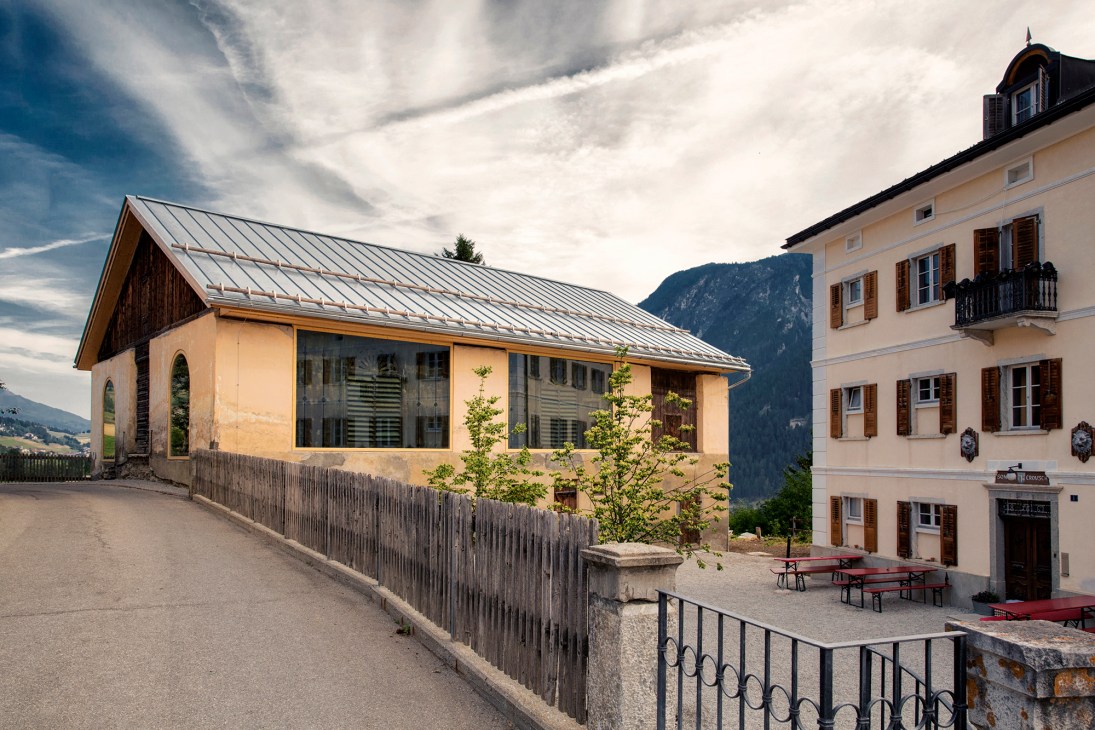 Modern timber building with metal roof and large windows next to traditional Alpine building with mountains behind