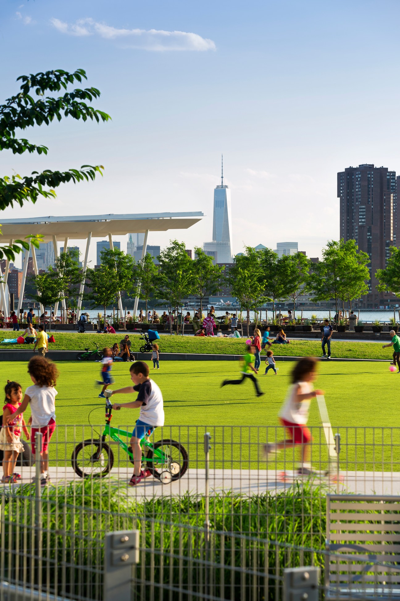 Children play on a grass oval in a waterfront park with One World Trade Center visible in the background.