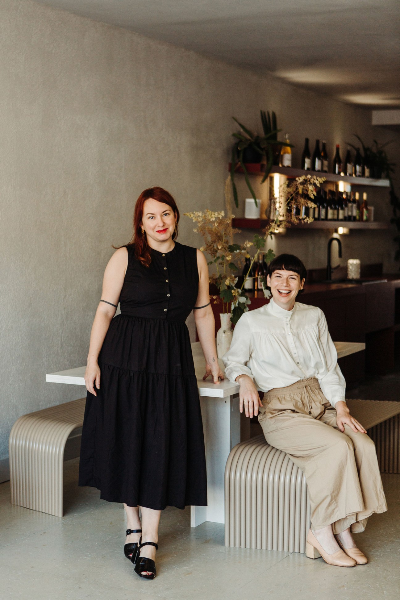 Two women at Grape Glass wine shop in Toronto, one standing in black dress, one seated in beige outfit