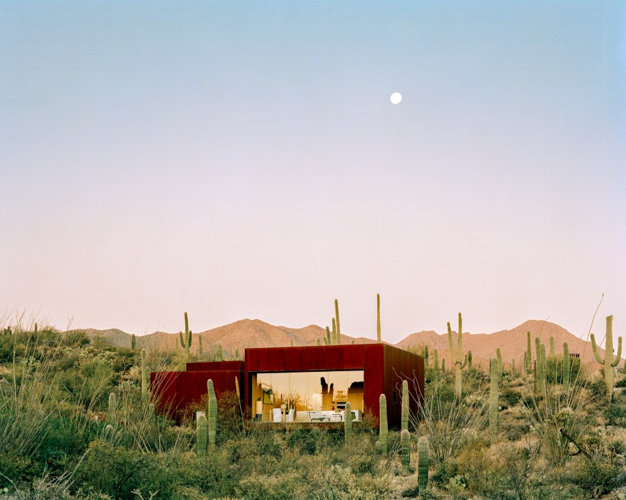 Modern red concrete house integrated into Arizona desert landscape with saguaro cacti and mountains at dusk