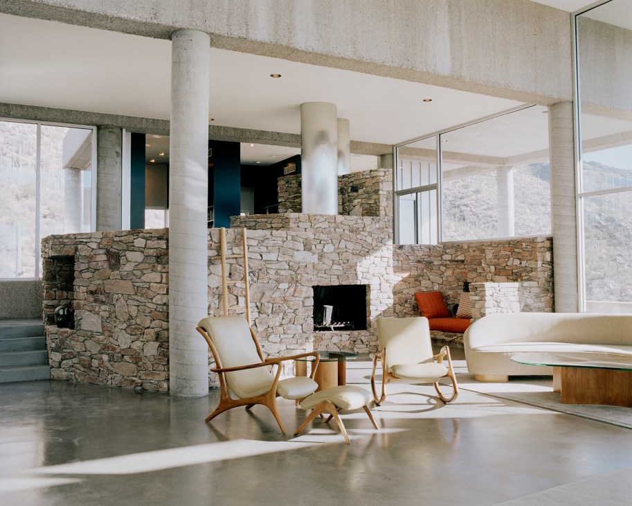 Modernist living area with stone walls, concrete columns, and mid-century furniture overlooking desert landscape