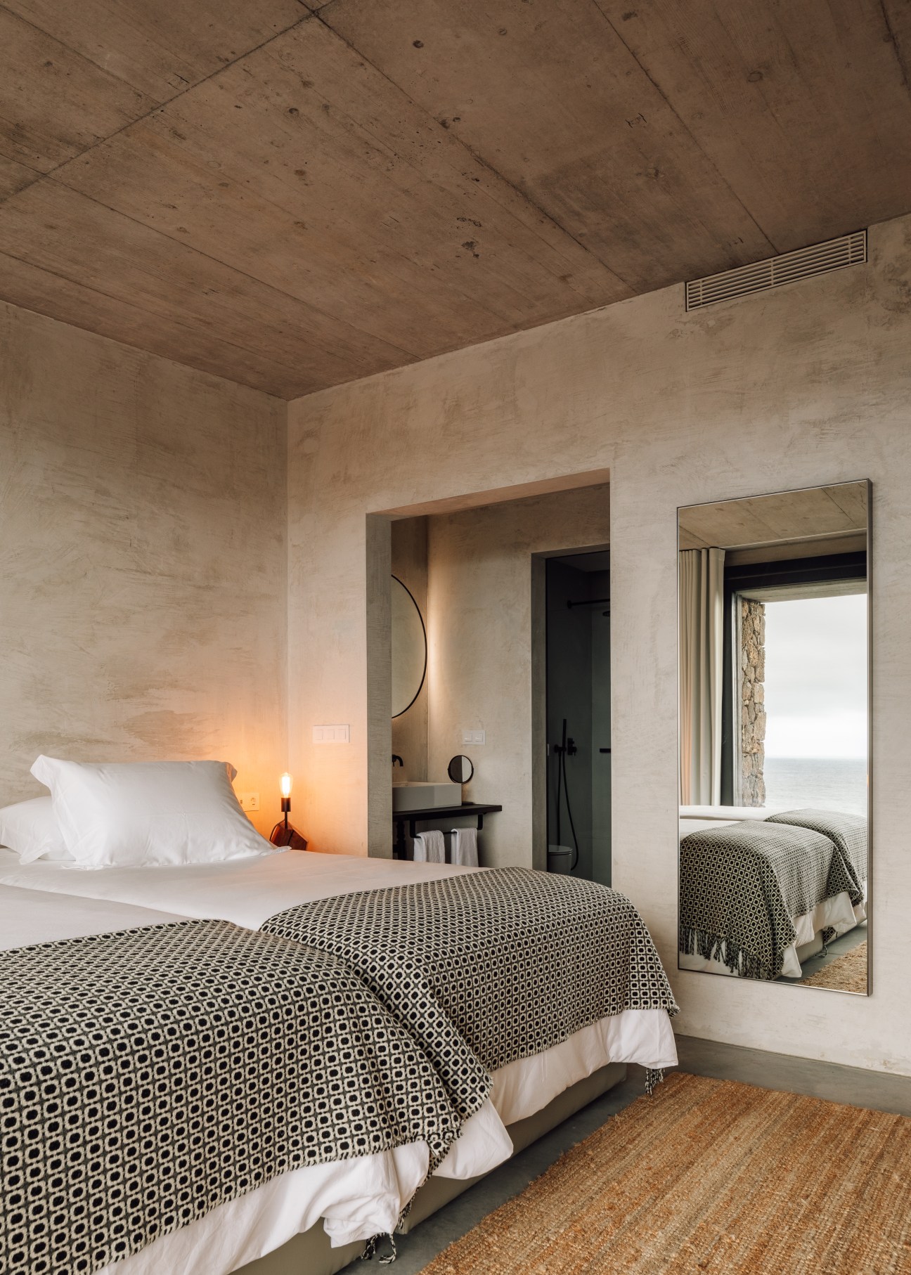 Minimalist apartment bedroom with charred wood ceiling, geometric patterned bedcover, white pillows, and vanity alcove with round mirror