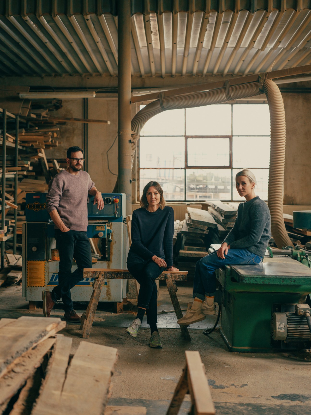 Three people stand in a wood workshop with machinery and large windows overlooking a city.