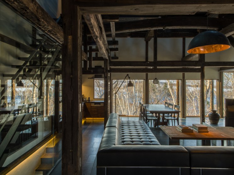 Modern living space with exposed wooden beams, black pendant lamp, and floor-to-ceiling windows overlooking bare trees at Shiguchi in Hokkaido.
