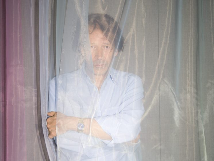 Portrait of designer Peter Saville behind sheer curtains wearing a light blue shirt