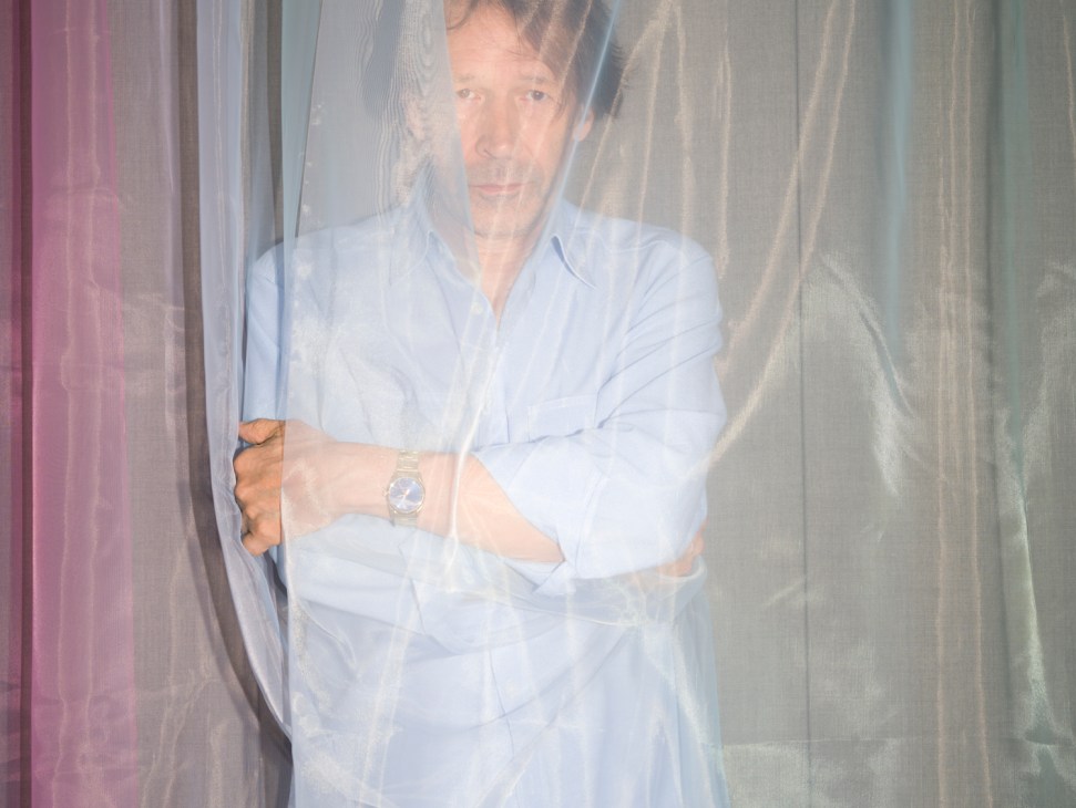 Portrait of designer Peter Saville behind sheer curtains wearing a light blue shirt