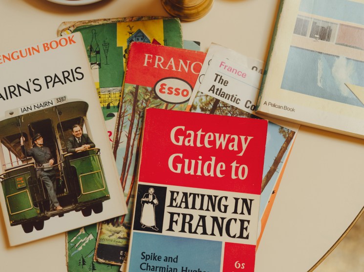 Collection of vintage travel guidebooks including Gateway Guide to Eating in France spread on table with coffee cup