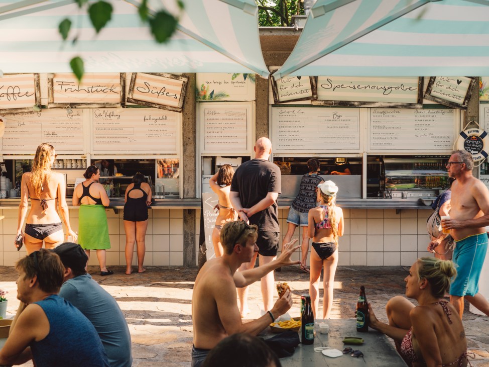 People in swimwear ordering food from outdoor food stalls under striped umbrellas