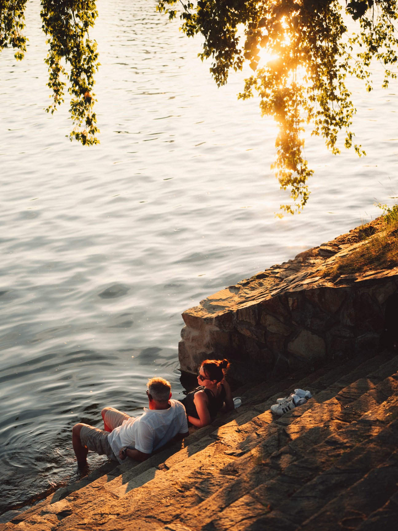 Two people sitting on rocky waterfront ledge at sunset with overhanging tree branches