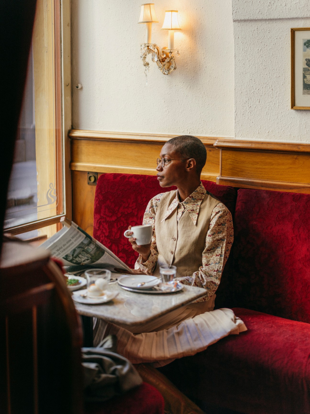 Woman in patterned shirt reading newspaper while having coffee at cafe table with red banquette seating