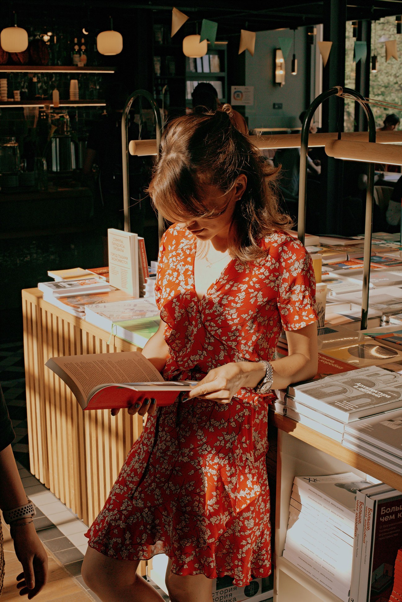 Woman in floral dress reading book in modern bookshop with wooden fixtures and stacked books