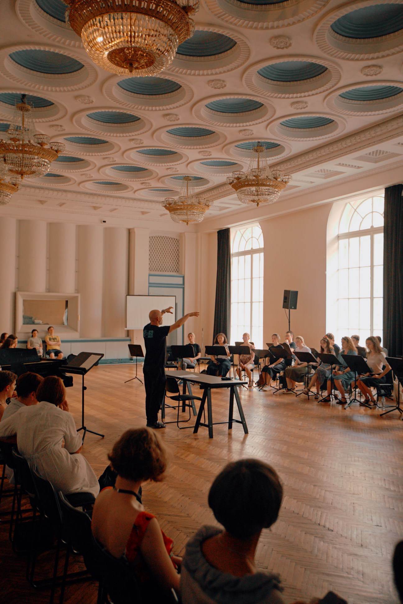 Conductor leading choral masterclass for students in ornate room with decorative ceiling and crystal chandeliers