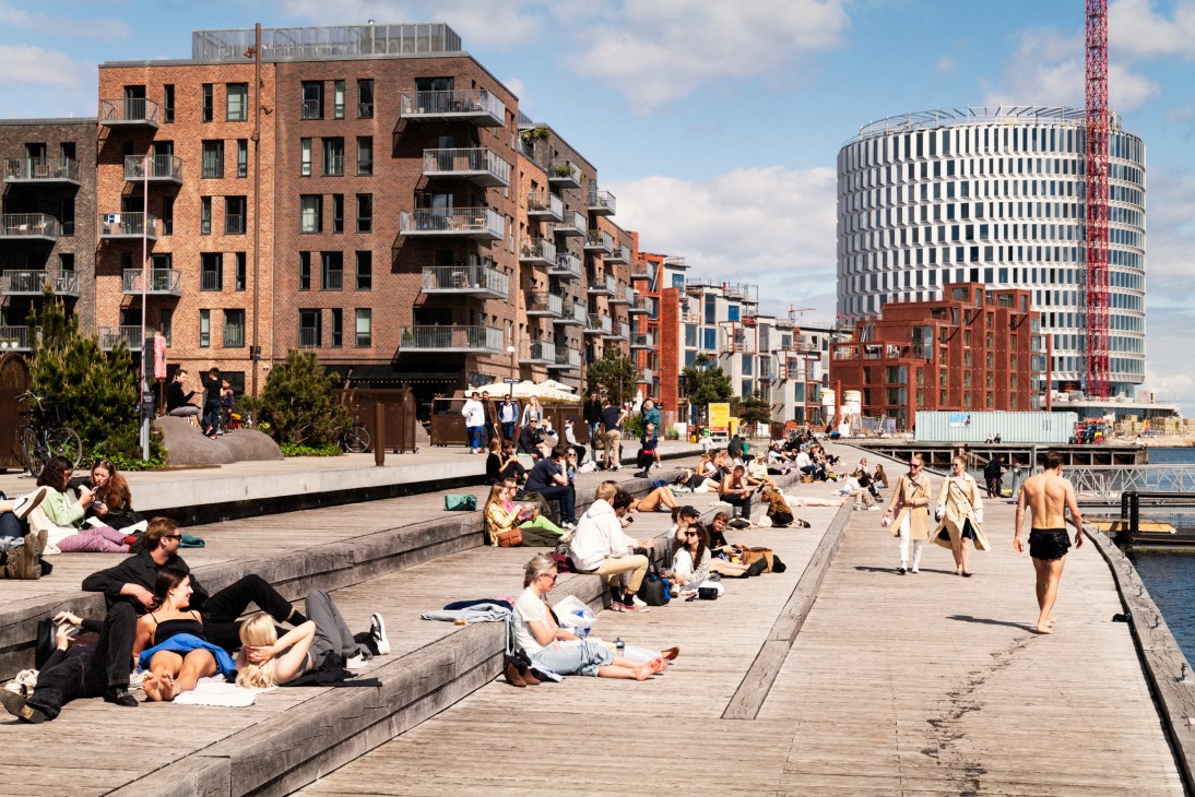People sunbathing and relaxing on wooden waterfront boardwalk with modern apartment buildings and harbor