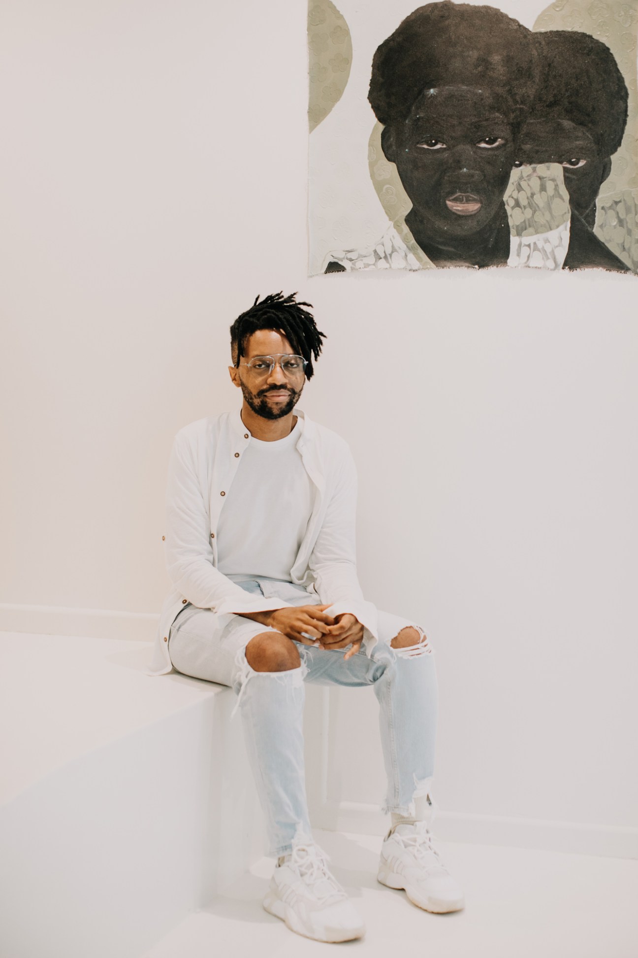 Artist Banele Khoza sitting on floor in white clothing in front of artwork