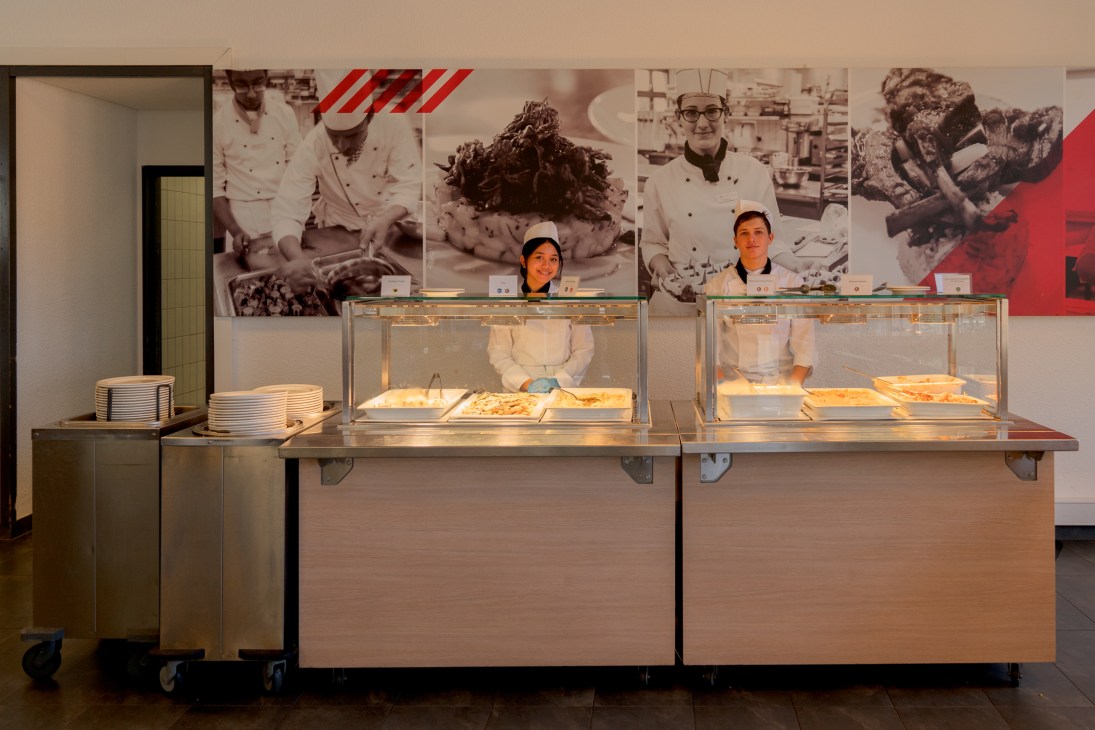 Two students in white chef uniforms and caps working behind glass display cases in a professional kitchen