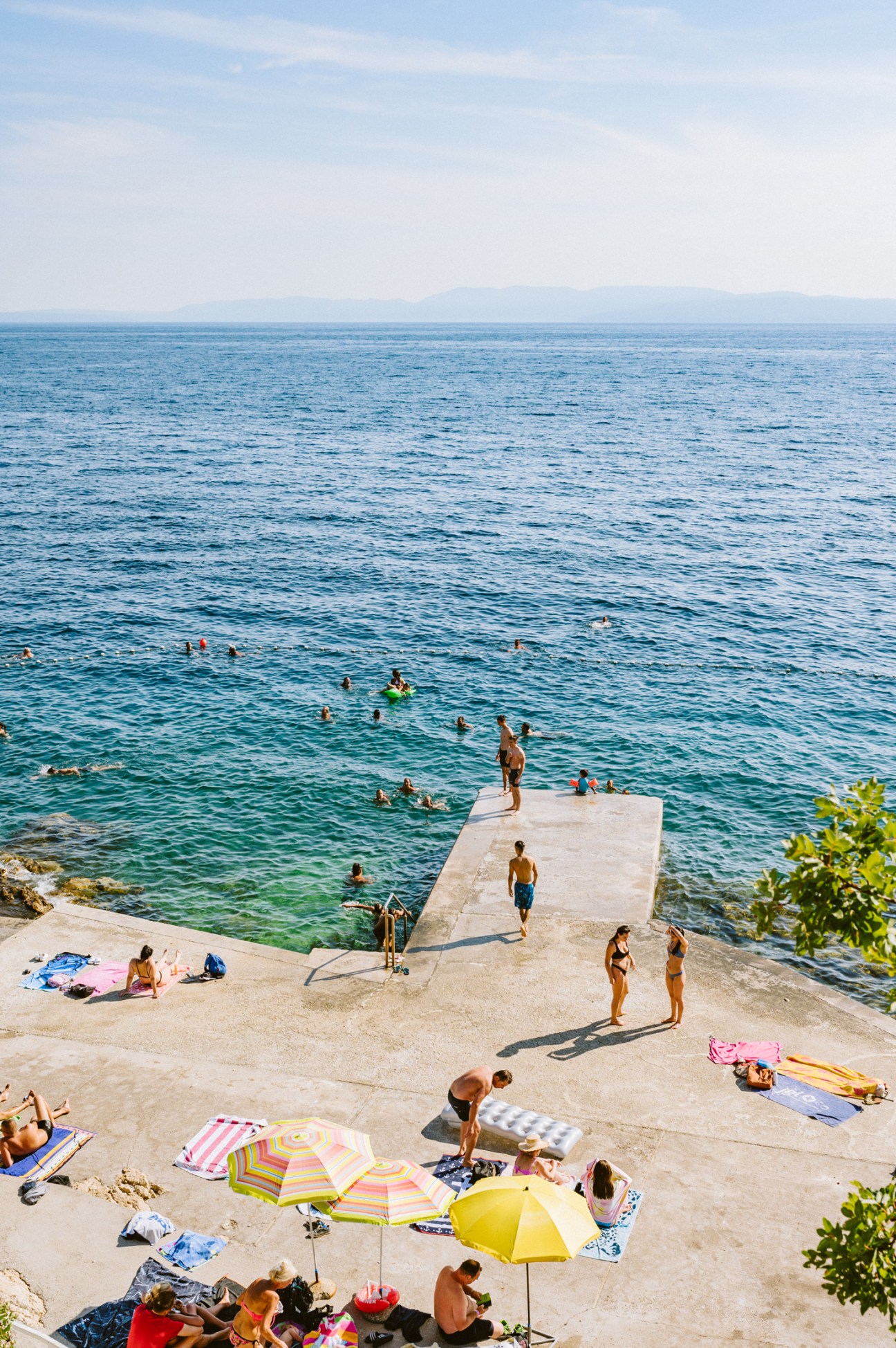 People swimming and sunbathing at a concrete waterfront area with colorful umbrellas and towels by blue ocean water