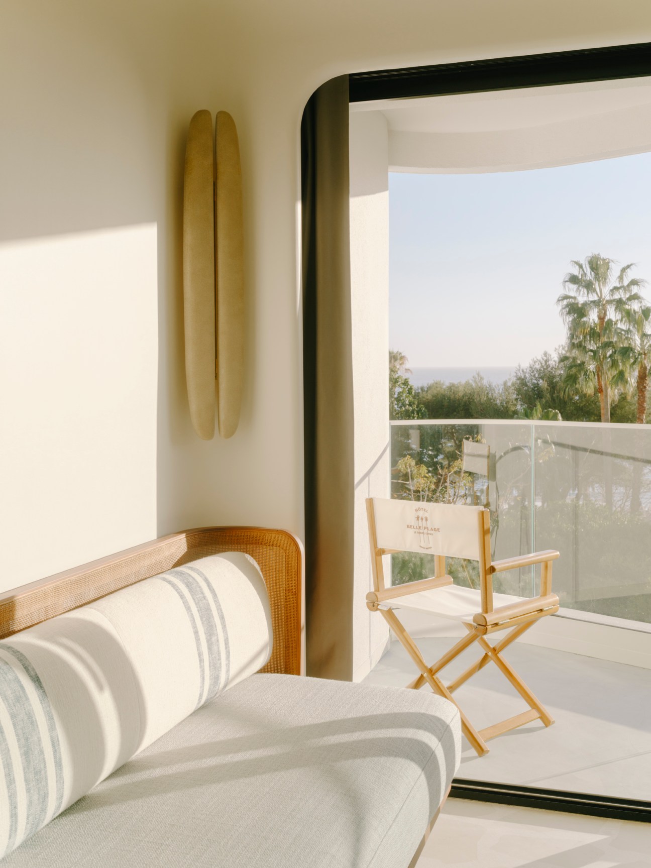 Modern hotel room interior with cane-backed chair, striped upholstered sofa and balcony overlooking palm trees