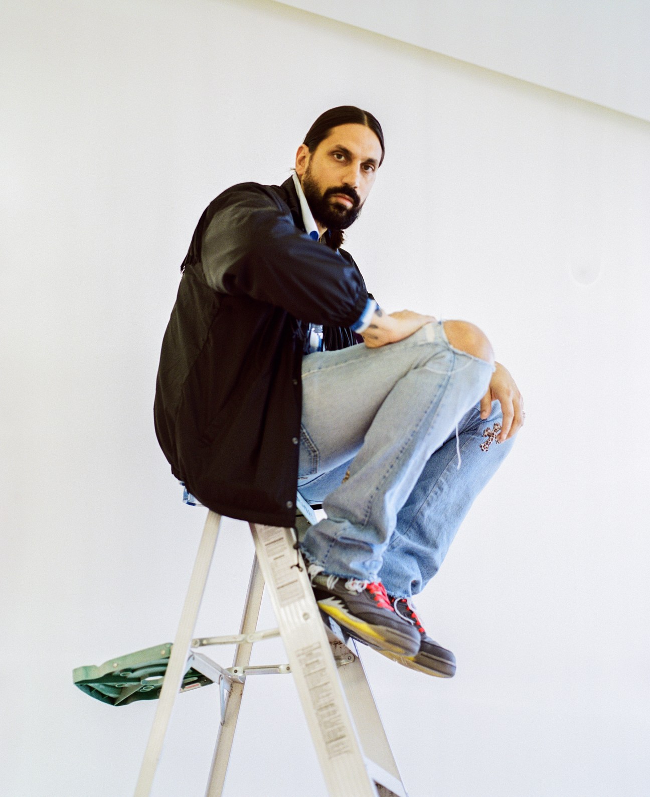 Ben Gorham, founder of Byredo, sits on a ladder wearing a black jacket and distressed jeans.