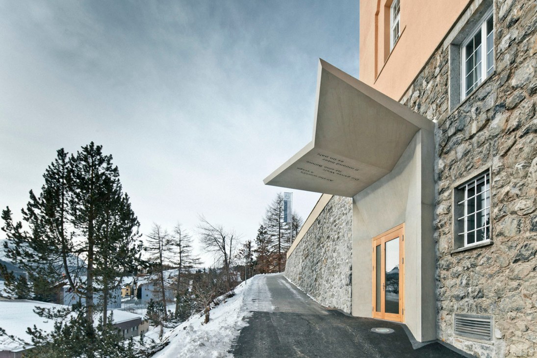 Modern angular building with distinctive overhanging roof element and illuminated entrance in snowy Alpine setting