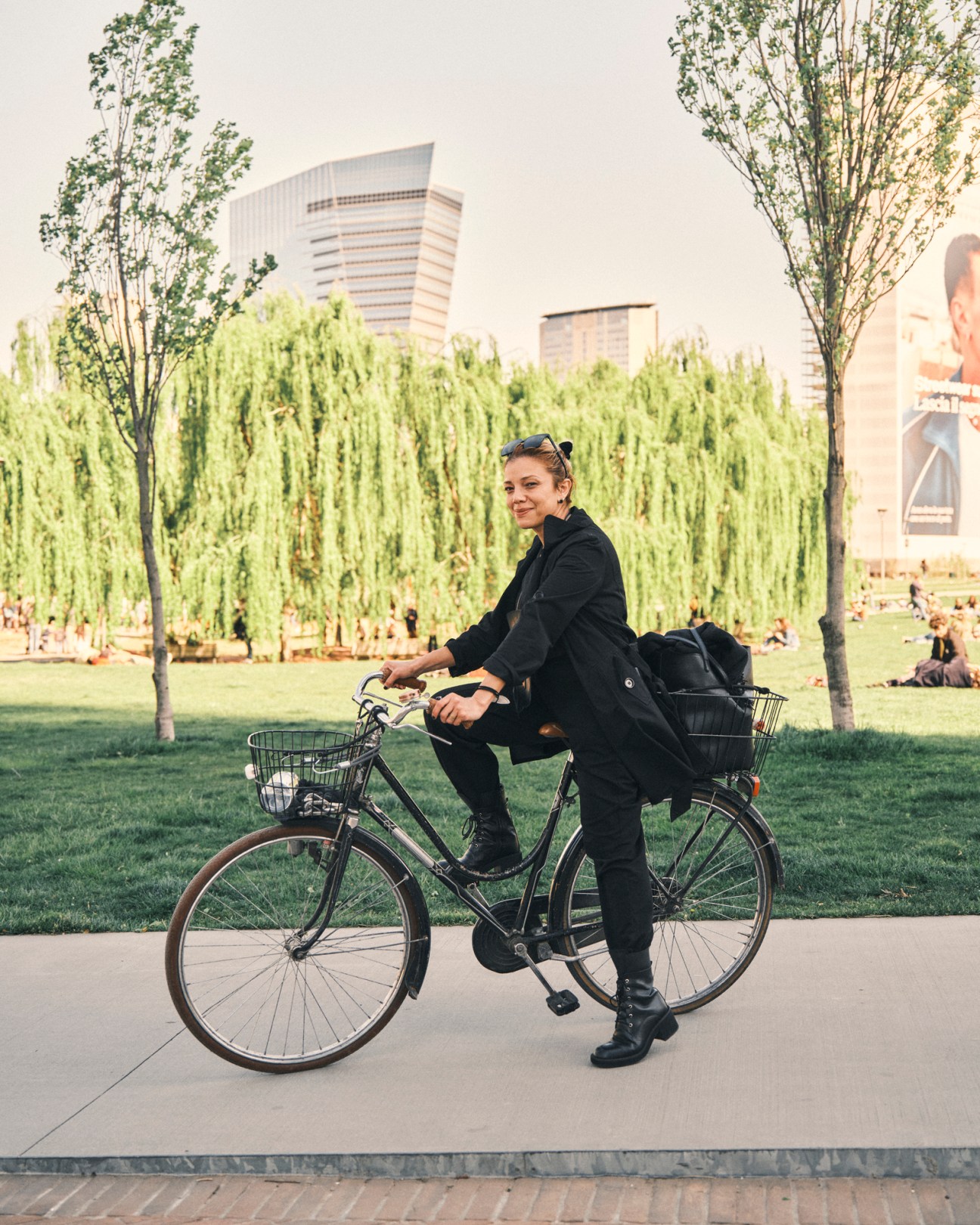 Woman in black jacket sitting on bicycle with front and rear baskets on urban path with park and modern buildings behind